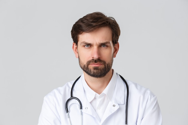 Hospital, healthcare workers, covid-19 treatment concept. Close-up of serious, determined bearded doctor in white scrubs and stethoscope, frowning looking camera, listening to patient.