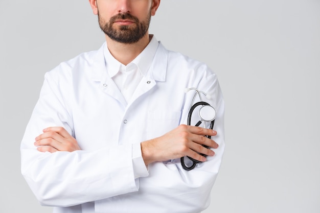 Hospital, healthcare workers, covid-19 treatment concept. Cropped shot of serious-looking confident doctor with beard, wearing white scrubs. Physician cross arms chest, holding stethoscope.