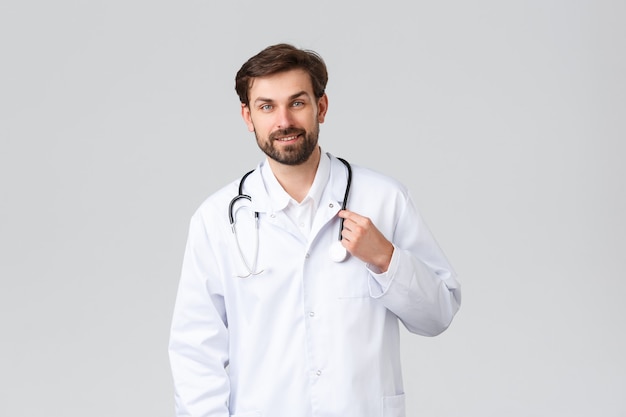 Hospital, healthcare workers, covid-19 treatment concept. Handsome bearded doctor in white scrubs with stethoscope, smiling at patient, listening to problems, treating people at clinic