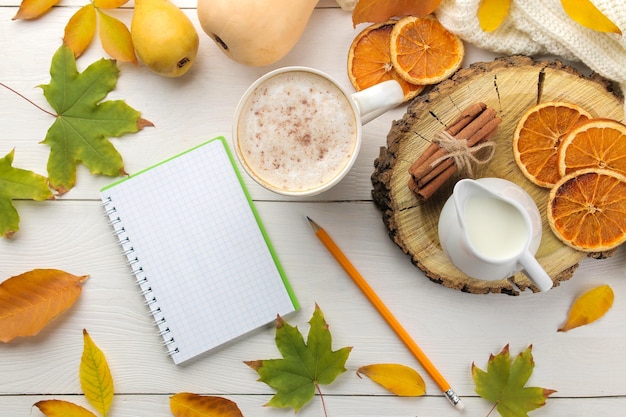 Hot autumn drink coffee or cocoa with a notebook yellow leaves and decorative pumpkins on a white wooden table autumn composition top view