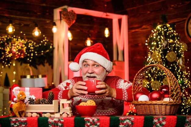 Hot beverage. Christmas cocoa recipe. Garland lights cozy atmosphere. Cozy home. Senior man Santa claus drinking tea. New year. Grandpa with mug. Christmas eve treats. Cozy interior. Winter holiday.