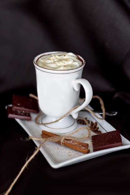 Hot chocolate in a mug with handle, spices and chocolate pieces on a white plate on a black background