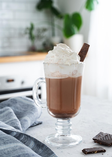 Hot chocolate with whipped cream and chocolate pieces in a tall glass with a cinnamon stick on the background of a white kitchen in the early morning Front view and close up