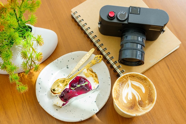 Hot coffee latte with latte art milk foam in cup made of paper and camera in notebook Blueberry Cake on wood desk on top view As breakfast In a coffee shop at the cafeduring business work concept