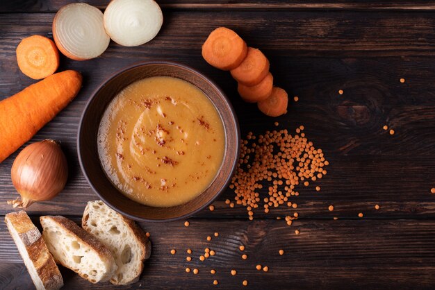 Hot delicious red lentil cream soup with ingredients: carrot, onion, and bread on a wooden table, close up, top view.