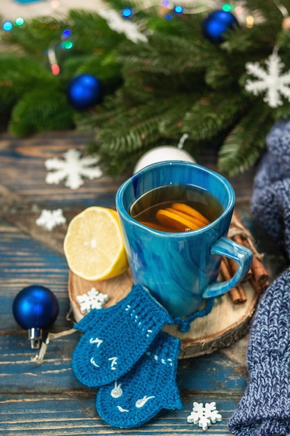 Hot drink with crocheted funny mittens. Traditional winter decor, fir tree branches, snowflakes. Festive seasonal arrangement in blue tones, old wooden background, close up