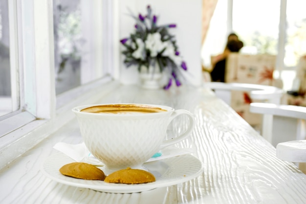 Hot latte coffee in a white coffee cup with cookies on white wooden table.