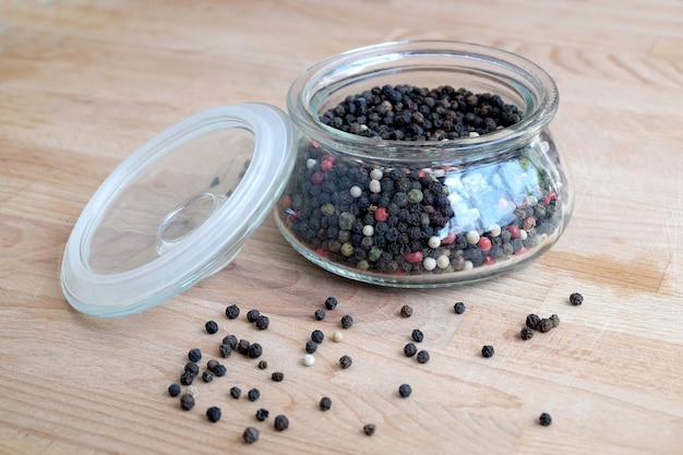 Hot spice ingredients for food. Still life with black pepper seeds inside round glass jar and scattered on wooden table