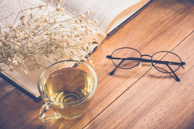 Hot tea in a cup with reading glasses on old book and flower on table