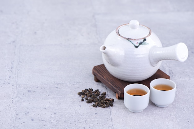 Hot tea in white teapot and cups on a sieve over bright gray cement background, closeup, copy space design concept.