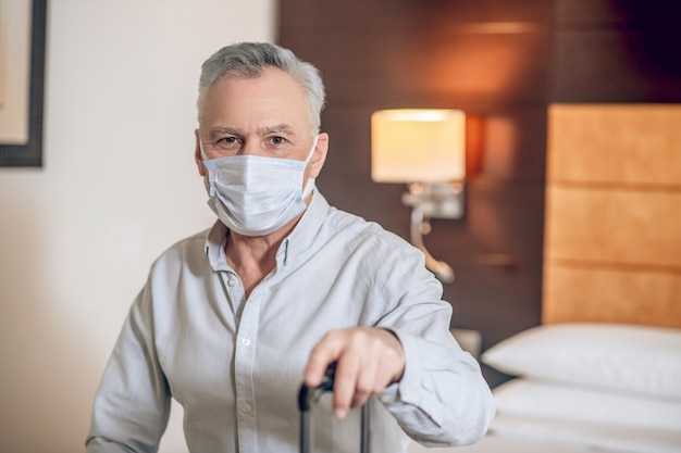 In the hotel room. Gray-haired mid-aged man with a protective mask in a hotel room