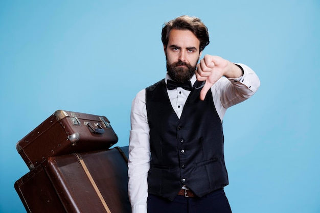 Hotel staff seems unsatisfied and unhappy while giving thumbs down on camera, dressed in suit and tie. Skilled bellhop indicates negative emotions of refusal and disappointment that lead to sadness.