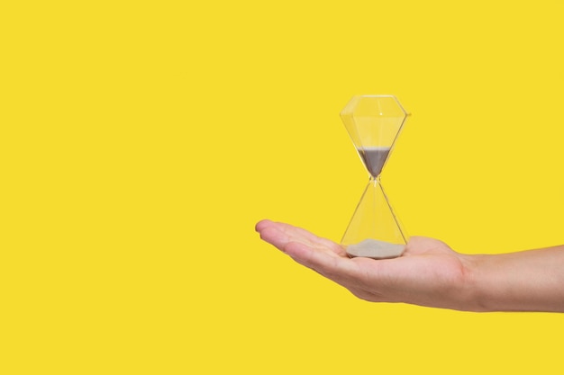 An hourglass on palm of your hand, yellow background, concept of loss and old age passing time.