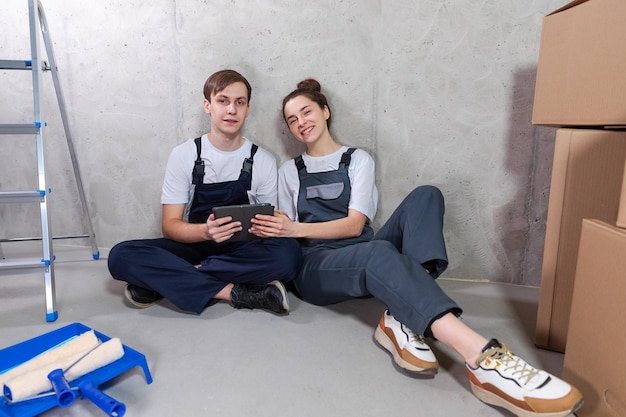 House DIY renovation concept Young happy couple sitting on floor looking home repair plan in tablet indoors Woman showing to man plan interior design online Couple in new home during repair works