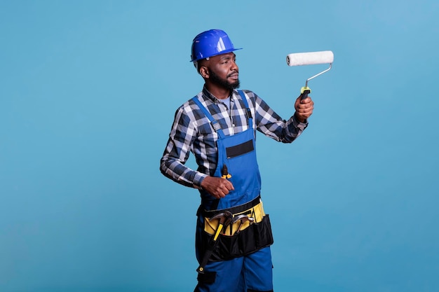 House painter making interior renovation, painting walls with roller. Construction worker wearing uniform and protective helmet isolated on blue background, studio shot.