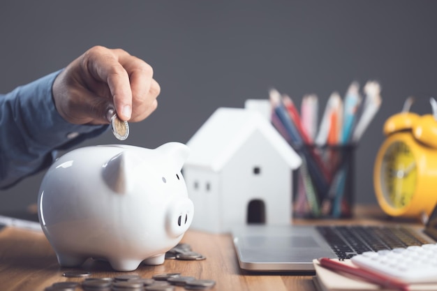 House purchase and money saving idea Home cost calculate concept Man adding coin to white piggy bank on office desk to plan for house financial Residence buying planning in the future