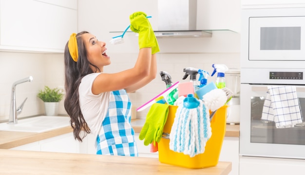 Housekeeping young lady with a yellow bucket full of cleaning stuff on her kitchen desk doing frolics with a kitchen brush pretending to sing into it.