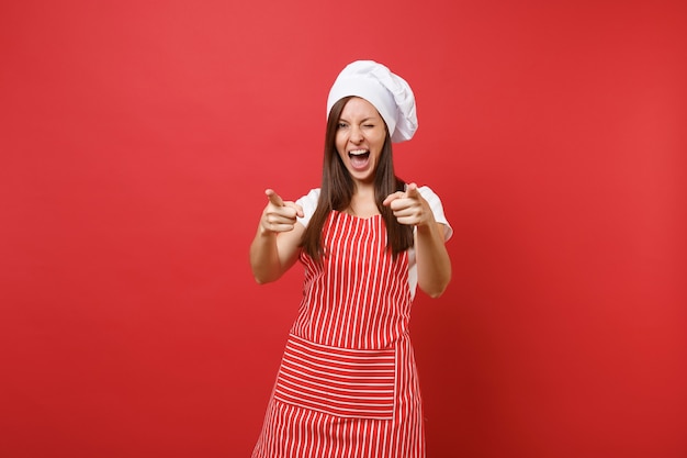 Housewife female chef cook or baker in striped apron, white t-shirt, toque chefs hat isolated on red wall background. Beautiful housekeeper woman pointing fingers camera. Mock up copy space concept.