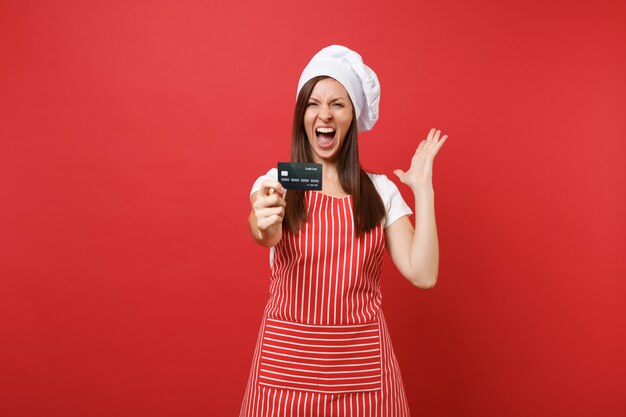 Housewife female chef cook baker in striped apron white t-shirt toque chefs hat isolated on red wall background. Excited woman hold in hand credit bank card, cashless money. Mock up copy space concept