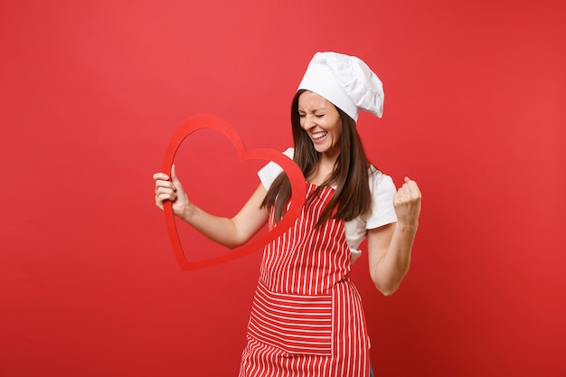 Photo housewife female chef cook or baker in striped apron, white t-shirt, toque chefs hat isolated on red wall background. smiling housekeeper woman holding wooden red heart. mock up copy space concept.