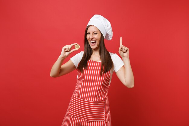 Housewife female chef cook or baker in striped apron white t-shirt toque chefs hat isolated on red wall background. Smiling woman holding bitcoin, bit coin future currency. Mock up copy space concept.