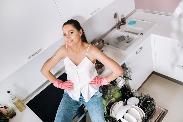 A housewife girl in pink gloves after cleaning the house sits tired in the kitchen.In the white kitchen, the girl has washed the dishes and is resting.Lots of washed dishes