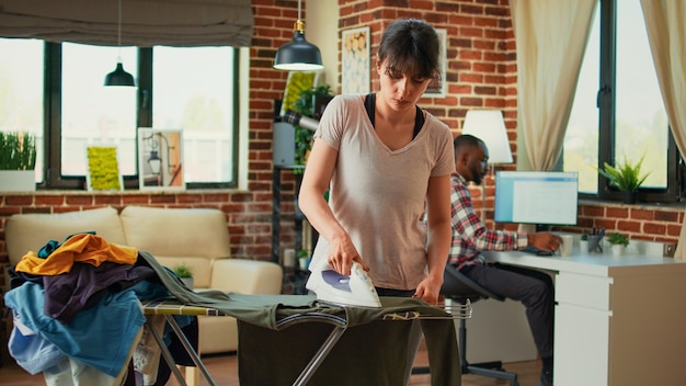 Housewife using iron with steam to smooth out clothes after washing them, ironing pile of laundry on ironing board. Casual person doing spring cleaning at home, domestic lifestyle.