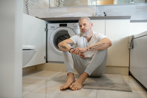 Housework. A gray-haired man sitting on the floor in the bathroom and looking upset