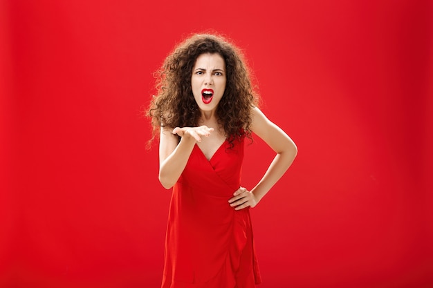 How dare you betray me. Pissed outraged bossy and displeased european adult woman with curly hairstyle in elegant dress bending towards camera with raised palm, complaining blaming and judging.