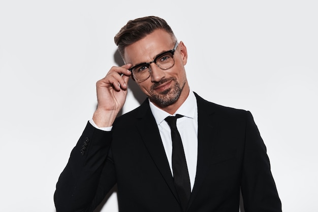 How may I help you? Handsome young man in full suit adjusting eyewear and looking at camera with smile while standing against white background