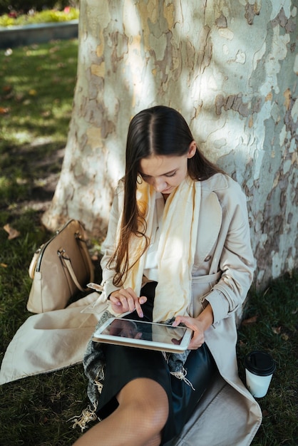 Under a huge plane tree in the park a girl sits with a tablet pc in her hands and writes something with her finger on the screen