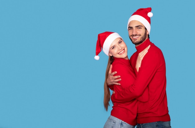 Photo hugging young couple in santa hats posing over blue background in studio