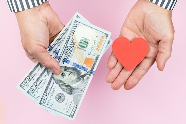 Human hands holds red heart in one hand and several hundred dollar bills in the other hand