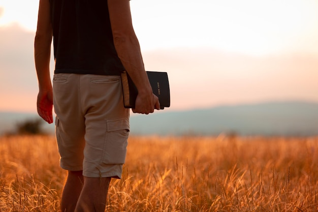 Human praying on the holy bible in a field during beautiful sunset.