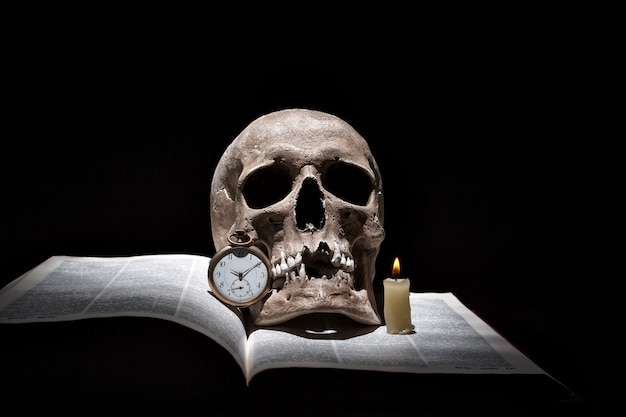 Human skull on old open book with burning candle and vintage clock on black background under beam of light