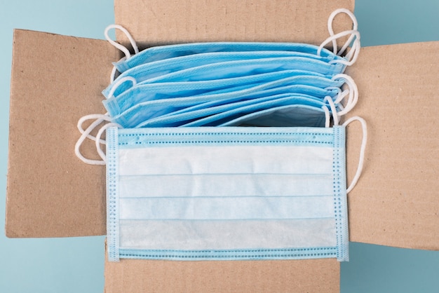 Humanitarian aid help concept. Top above overhead close up view photo of open unpacked unwrapped paper box with lot of medical masks on blue background