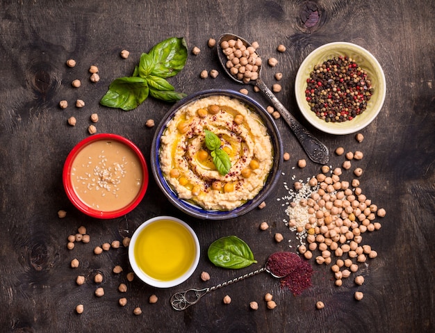 Hummus ingredients. Chickpea, tahini paste, olive oil, sesame seeds, sumac, herbs on dark rustic wooden background.