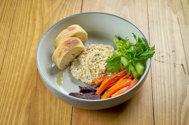 Hummus with baked vegetables and baguette in a bowl on a wooden background. Vegetarian dish. Israeli cuisine