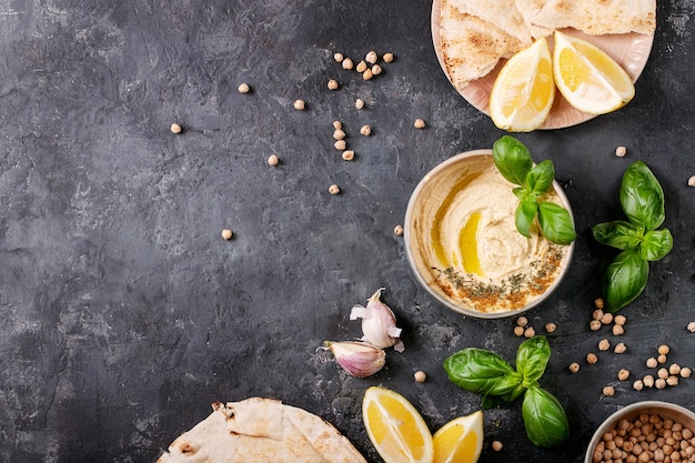 Hummus with olive oil, pita bread and ground cumin in ceramic bowl served with lemons, basil and chickpeas over dark texture surface. Top view, flat lay