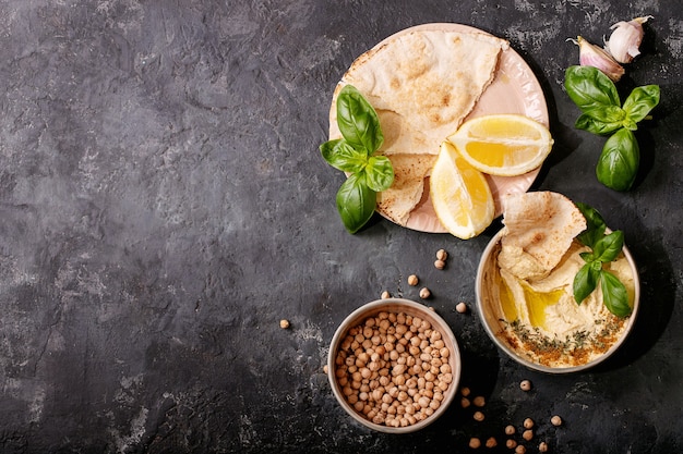 Hummus with olive oil, pita bread and ground cumin in ceramic bowl served with lemons, basil and chickpeas over dark texture surface. Top view, flat lay