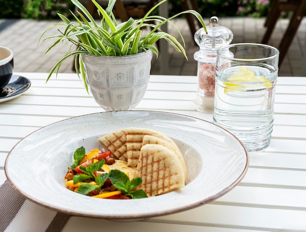 Hummus with tomatoes and herbs in white plate with pita on table and glass of water cafe vegan food