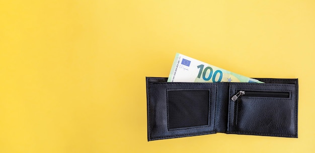 A hundred euro banknote sticks out of a black wallet on a yellow background. cash paper currency