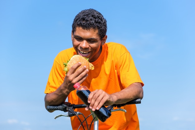 Hungry African American man enjoying the taste of hamburger and cola drink while sitting on bike outdoor in summer. Concept of love to junk food diet