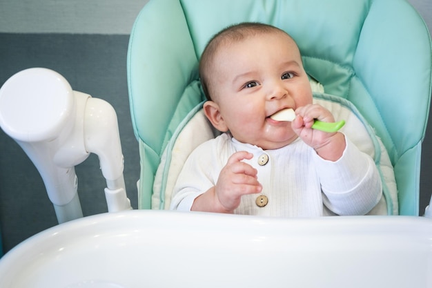 A hungry baby is gnawing on a plastic spoon at the table on a high chair Teething whims itchy gums introduction of complementary foods