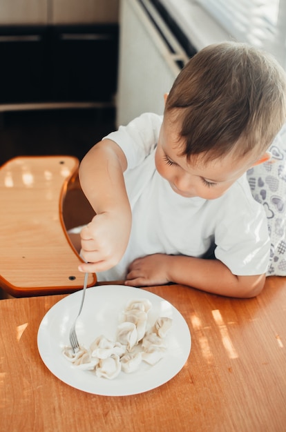 A hungry child is eating dumplings in the kitchen sitting in a chair in a white t-shirt