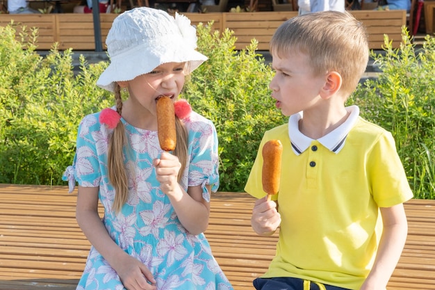 Hungry children eating street food sitting on a park bench A boy and a girl eating fried sausages on a stick