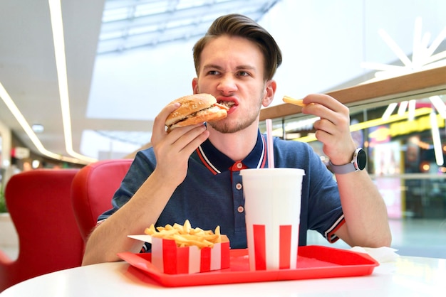 Hungry crazy man is biting eating fast junk food burger, French fries and drink soda. Hunger