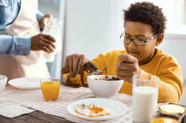 Hungry dinosaur. Cheerful pre-teen boy feeding a spoonful of cereals to a toy dinosaur while having breakfast