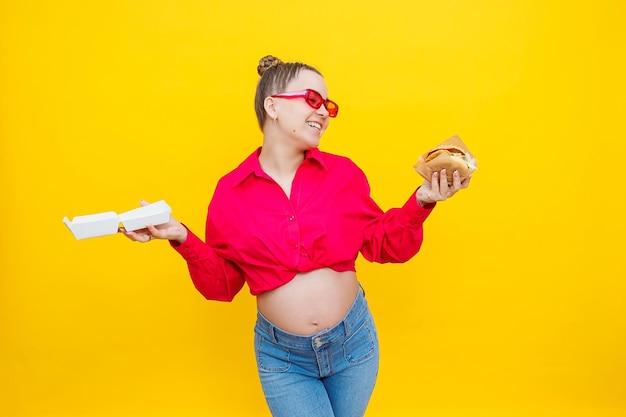 Hungry pregnant woman holding hamburger eating junk food posing on yellow background in studio Woman enjoying a big hamburger The concept of unhealthy eating and overeating during pregnancy