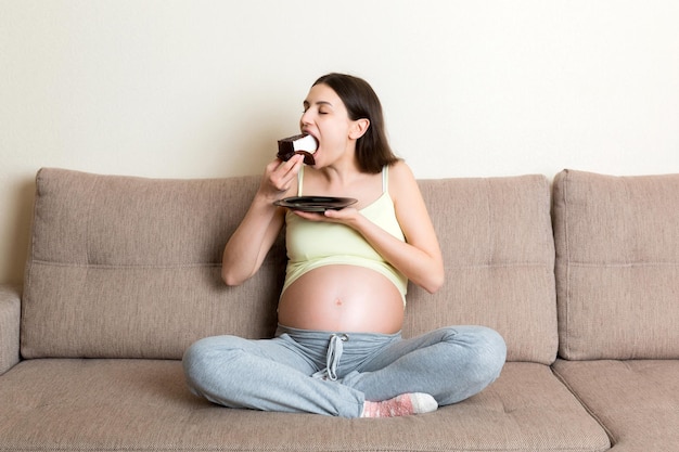 Hungry pregnant woman is eating a piece of tasty cake relaxing on the sofa at home Sweet cravings during pregnancy